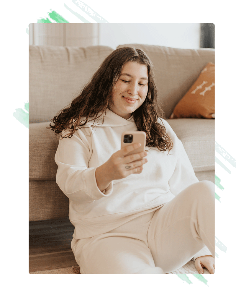 A girl sits in front of her couch looking at her phone and smiling.